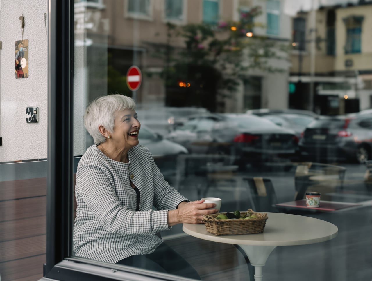 Candid documentary photo of an ordinary-looking woman in her sixties with short silver hair, sitting at a small window t